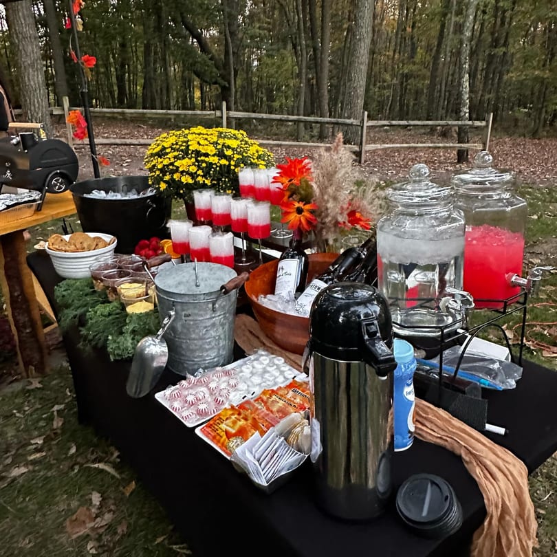Outdoor buffet table with drinks, snacks, and fall floral decorations in a wooded setting.