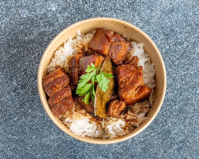 Top-down view of a takeout bowl filled with white rice, braised pork belly, bay leaves, and parsley.
