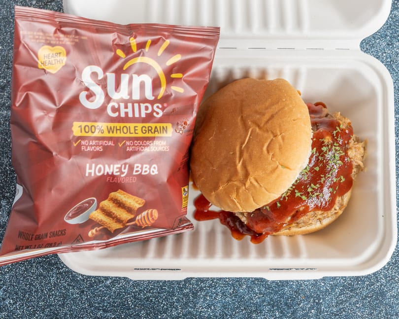 Pulled pork sandwich with BBQ sauce and a bag of Honey BBQ Sun Chips in a to-go container.