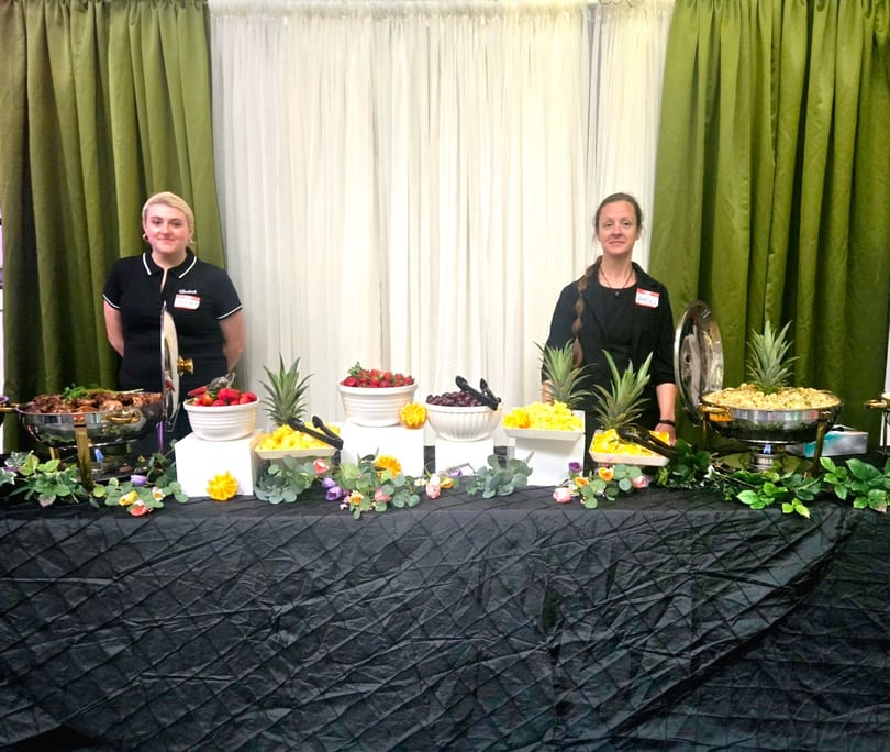 Two women stand behind a buffet table laden with fruit, hot food, and decorative foliage.
