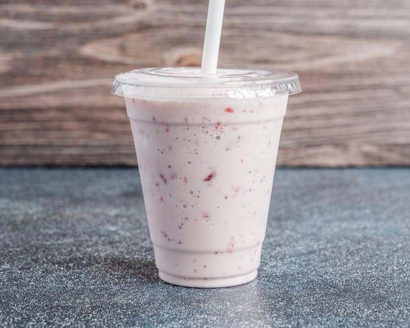 Strawberry smoothie in a clear plastic cup with a lid and white straw, against a wooden background.