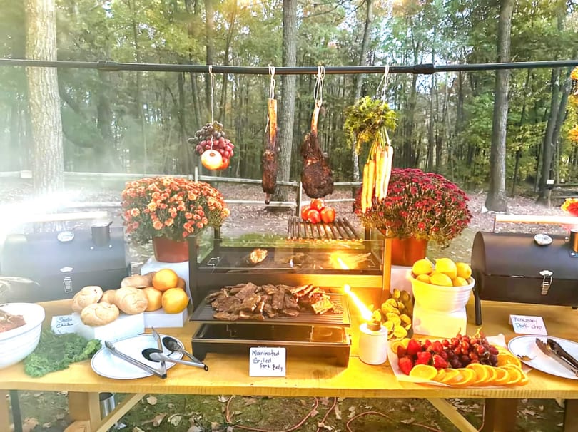Outdoor buffet table with grilled meats, fresh fruit, and vegetables, set against a forest backdrop.