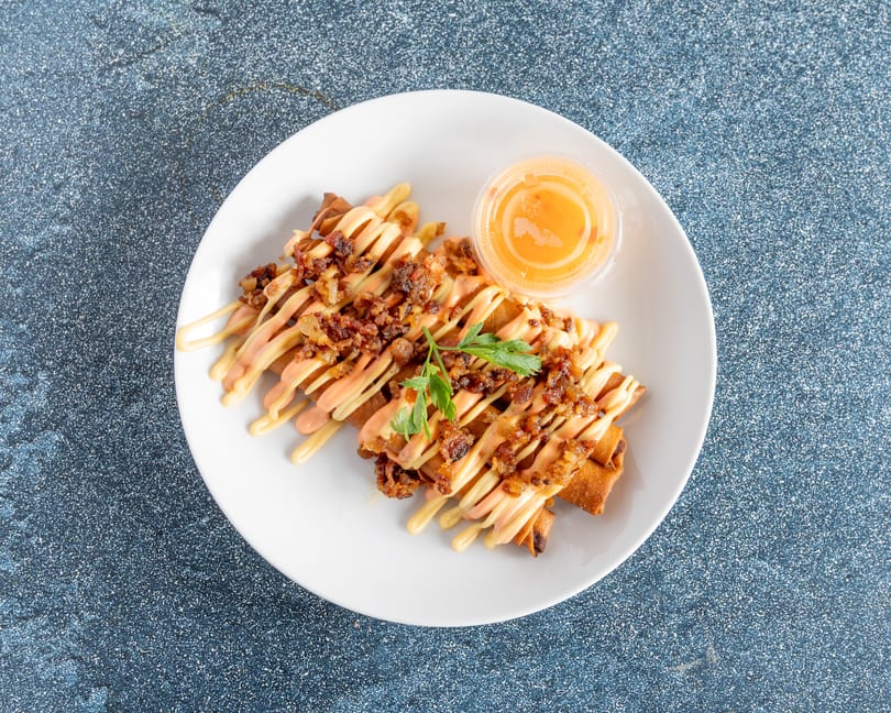 Crispy egg rolls drizzled with two sauces, topped with bacon bits and parsley, with a side of dipping sauce.