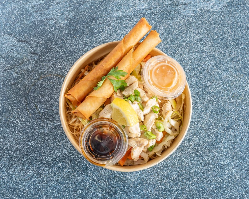 Top-down view of a noodle bowl with chicken, vegetables, two egg rolls, and sauces.
