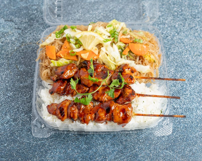 Chicken skewers with rice and pancit noodles, garnished with parsley and lemon, in a takeout container.